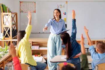 Professional friendly female teacher asking pupils while kids raising their hands up during lesson, classroom at private school