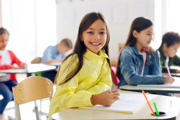 Happy school girl smiling at camera, sitting at desk in classroom, writing in notebook. Group of diverse classmates studying on background