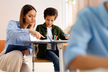 Professional female teacher helping with task to Latin schoolboy, pupils sitting at desks, studying in secondary school classroom