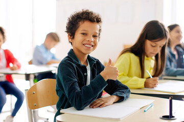Excited Latin schoolboy sitting at desk in classroom, showing thumb up and smiling at camera, classmates on background