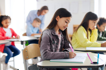 Study process concept. Pretty schoolgirl writing in copybook during lesson, having test, sitting at desk in modern classroom interior