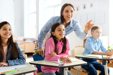Cheerful woman teacher explaining and pointing at blackboard while standing among pupils, creating an interactive learning atmosphere in classroom