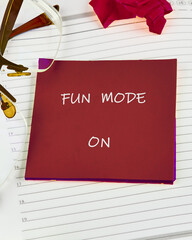 A red square paper with the inscription FUN MODE ON lies on a notebook, surrounded by glasses and a crumpled sheet of paper, suggesting a break from business for leisure