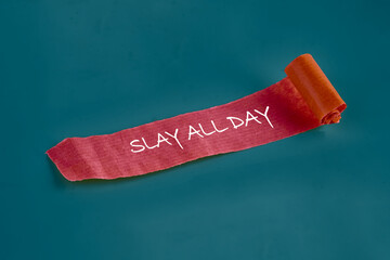 A red textured paper with white handwriting that says SLAY ALL DAY is partially unrolled against a Teal background in a studio setting, promoting confidence and goals