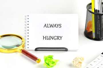 A notepad displays ALWAYS HUNGRY near a magnifying glass, crumpled paper, and office supplies, implying relentless pursuit of improvement and success in business