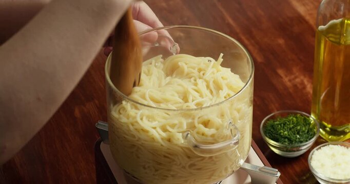 The cook stirs with a wooden spatula hot water boils in a glass saucepan with spaghetti inside, steam gently rising. Transparent cookware cooking pasta. 4K.