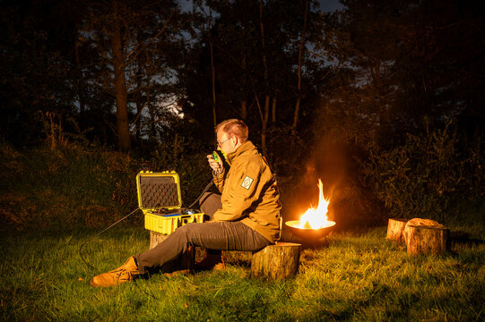 Amateur radio operator outdoors by a campfire, operating portable HF radio from a Go-Box in the wilderness, enjoying nature, emergency communication and Parks on the Air activity