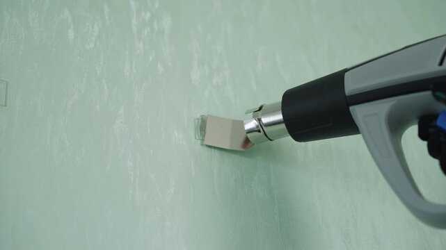 Close-up of handyman using heat gun to soften and remove old adhesive hook and residue from textured green wall, closeup diy renovation and repair task in home interior. Shooting in slow motion.
