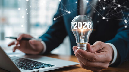 A person holding a lightbulb with the year 2026 written on it next to a laptop and a pen in hand