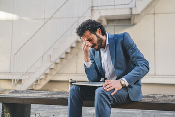 Stressed businessman sitting on bench with laptop is having headache