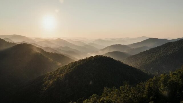 A serene landscape photograph showing layers of forested mountains under a soft, hazy sky with the sun