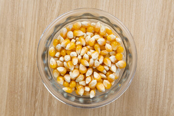 Yellow dried corn kernels in a glass vase in a light color wooden table. Close up and upper view.