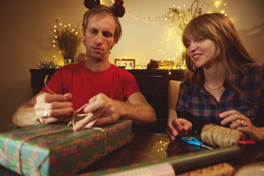 Couple Christmas Gift Wrapping At Cozy Dinner Table With Fairy Lights And Decorations For A Festive Home Moment - Powered by Adobe