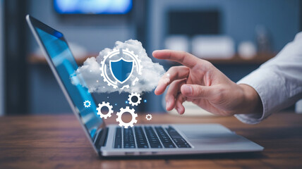 A laptop with a cloud and shield icon and a hand reaching for it on a wooden surface indoors