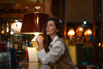 Young woman in white shirt and vest sitting at bar counter looking thoughtful. Warm light from...