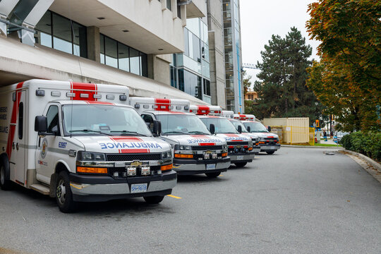 Row of white ambulances parked at modern hospital entrance under golden autumn trees, no parking sign for emergency vehicles