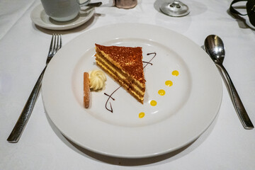 Elegant slice of classic Opera cake featuring delicate layers of coffee-soaked biscuit, ganache, and buttercream, dusted with cocoa powder and artfully plated with a cookie and cream swirl.