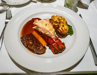 A surf and turf dish featuring a perfectly cooked steak, a succulent lobster tail, roasted carrots, and a side of golden-brown potato gratin, elegantly presented on a white plate.