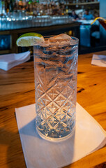 A refreshing gin and tonic or similar clear cocktail in a textured highball glass with ice and a lime wedge, resting on a napkin on a wooden bar top with a blurred background.