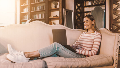 Fototapeta premium Portrait of casual cheerful lady using laptop sitting on sofa in living room and relaxing. Happy woman chatting with friends online, typing on pc keyboard, female freelancer working from home
