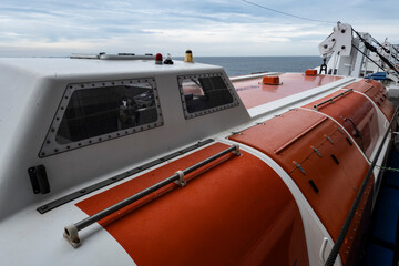 Atlantic Ocean - Sep 27, 2025 Close-up of an enclosed orange and white lifeboat on a ship’s deck with windows, hatches, and davits visible against a calm sea and cloudy sky.