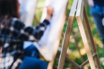 Fototapeta premium Kids drawing at plein air, group class of talented children with painting easel and wooden canvas during lesson of watercolor painting outdoors in summer, art school class for young teen artists