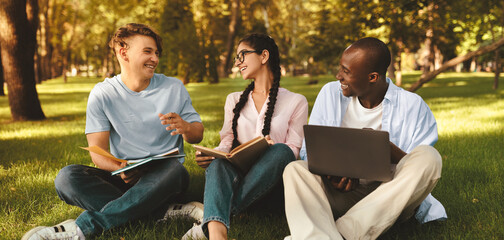 Happy international students resting in college campus, sitting on grass, talking and using laptop...