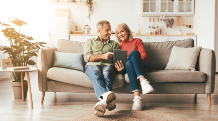 Cheerful Mature Spouses Using Digital Tablet Browsing Internet Or Watching Movie Together Sitting On Couch At Home. Senior People And Gadgets, Retirement Lifestyle Concept