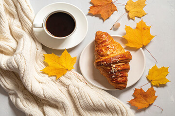Cozy morning flat lay with croissant, coffee cup, maple leaves, and knitted scarf &mdash; soft autumn breakfast mood.