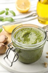 Tasty green pesto in glass jar and ingredients on white table, closeup