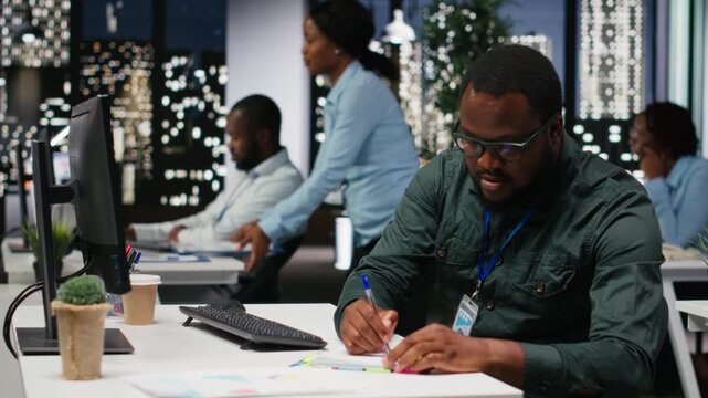 African american advisor extracting data by hand on a notebook late at night, drafting highlights from finance reports for the business administration. Notetaking supports goal setting. Camera B.