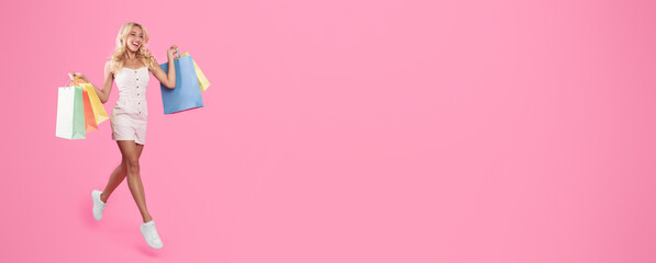 Seasonal Sales And Shopping Concept. Full length of happy blonde female consumer walking and jumping holding many colorful bags with new purchases isolated on pink studio wall, free copy space, advert