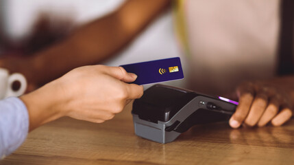 Cropped young black guy bartender in apron at bar counter gives terminal to woman client with credit card in cafe interior. Finance, order, work and small business, startup during covid-19 outbreak