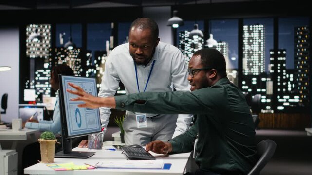African american workers team consulting each other in solving tasks at night, completing monthly reports and reviewing KPI data insights for productivity. Collaboration for success. Camera B.