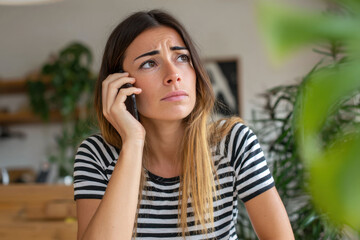 Fototapeta premium Portrait of a worried woman talking on the phone at home, showing concern or stress. Ideal for illustrating communication, anxiety, emotional situations, or modern lifestyle themes.