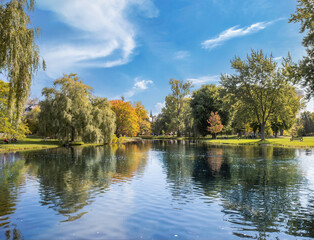 Looking across a lake at an urban park in autumn with willow and hardwood trees in fall colours, church steeple in background