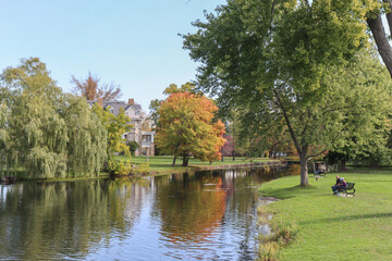 A small lake in an urban park in autumn, willow and hardwood trees in fall colours refected in water,  stone home in background, people on benches