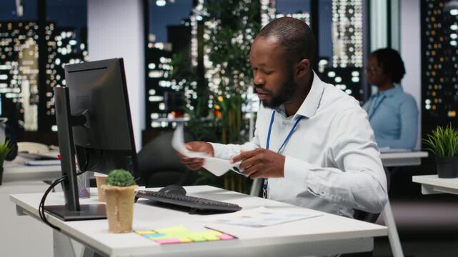 Black young employee proofreading financial data for a new report at night, reviewing archived files in the strategy room. Evaluating data, researching and organizing agenda. Camera B.