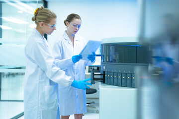 Two female laboratory scientists collaborate in a modern clinic. One loads an automated analyzer while her colleague verifies the procedure, ensuring accuracy in patient diagnostics.