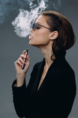 Obraz premium Young woman vaping with smoke in a dark studio, wearing sunglasses and black jacket, stylish and modern.