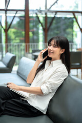 Fototapeta premium Asian woman freelancer talking on phone and holding pen with notebook while sitting on sofa in cafe