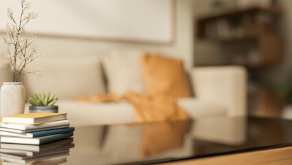 Books with flower vase and pot plant on glass top wooden table aside sofa in a blurred living room.
