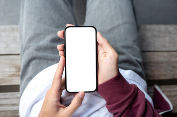 A man's hands holding blank screen smartphone in both hands while sitting outdoor on wooden bench.