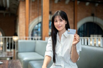 Pretty asian woman holding or showing a credit card while sitting on sofa in a cafe or coffee shop.