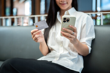 Close up of asian woman holding credit card and looking at smartphone while sitting on sofa in cafe.