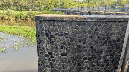 A sturdy dam wall made of textured river stones stands next to a reservoir bridge green foliage,...
