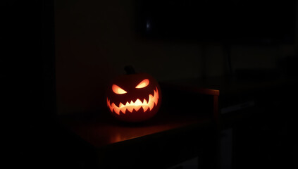 Photograph of a carved pumpkin with a sinister expression, glowing from within. The pumpkin has triangular eyes and a jagged, toothy smile, illuminated by an orange light that contrasts with the dark