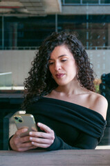 Argentinian woman with curly hair focused on her smartphone at an outdoor café. Neutral expression, relaxed and elegant atmosphere.