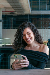 Argentinian woman with curly hair smiling while using her smartphone in an outdoor café. Bright and cheerful mood.