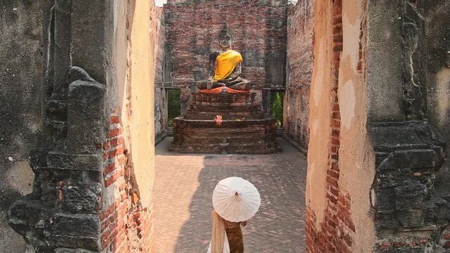Thailand, Ayutthaya - march 15th, 2023: Woman in traditional Thai dress holding umbrella sightseeing visit ancient Buddha statue.Ayutthaya historical park. Concept heritage, spirituality, thai culture
